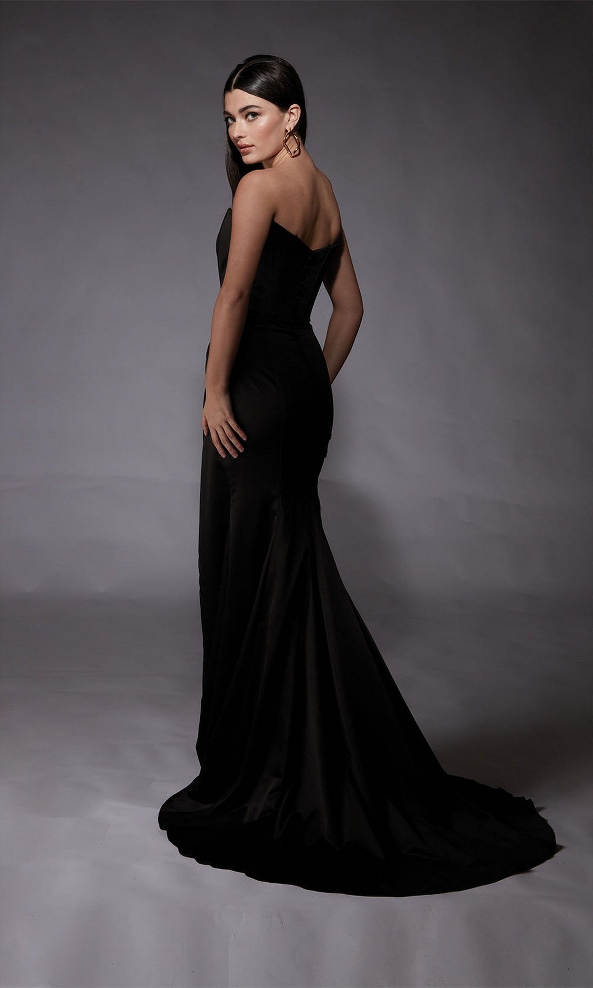 Woman modeling the Alyce 7093 long formal dress—a strapless satin gown with a fitted sheath silhouette and sweeping train—stands on a grey background, glancing over her shoulder.